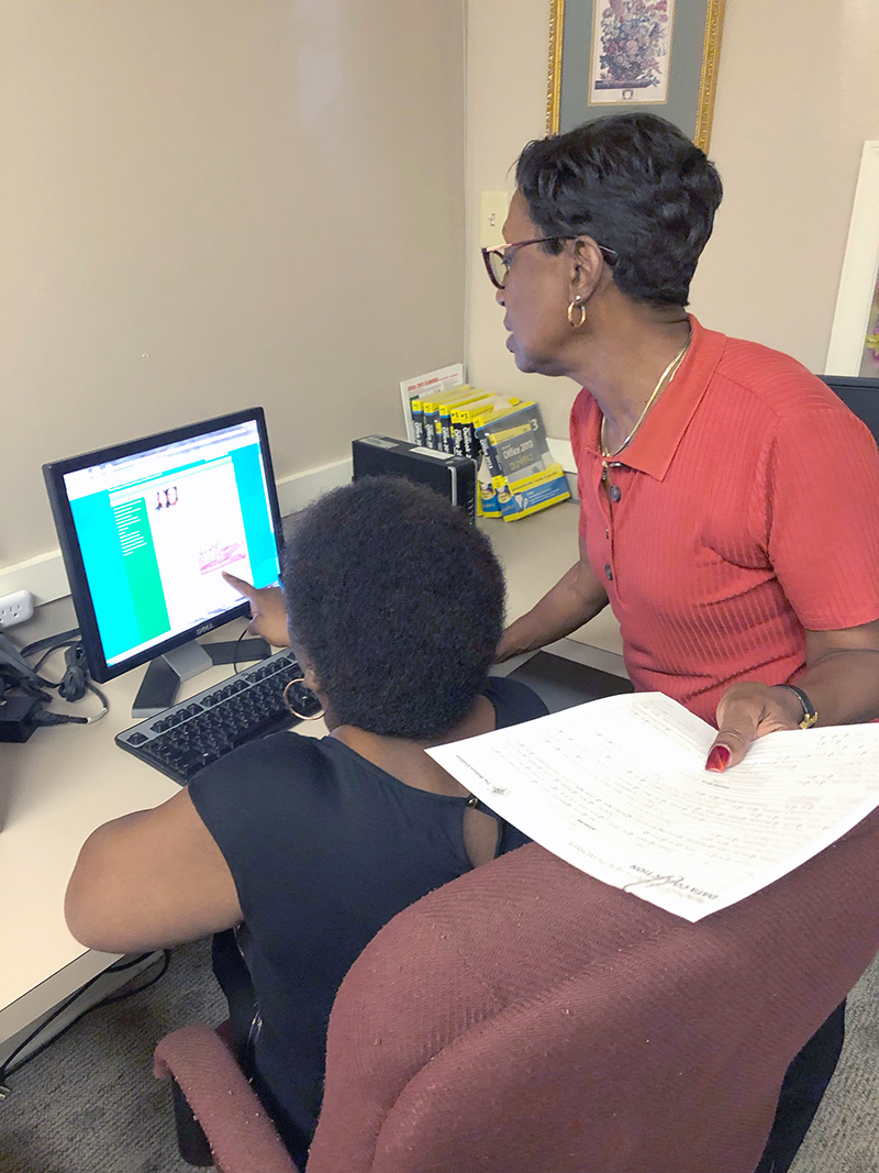 Pat Adams standing behind another woman at a computer screen showing her something